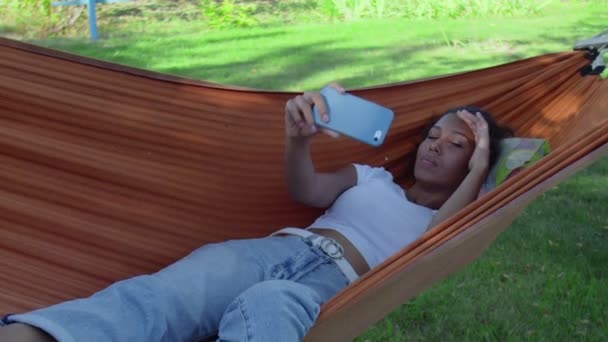 Młoda, szczęśliwa, afrykańska kobieta robi sobie selfie z telefonem leżącym na hamaku - Materiał filmowy, wideo
