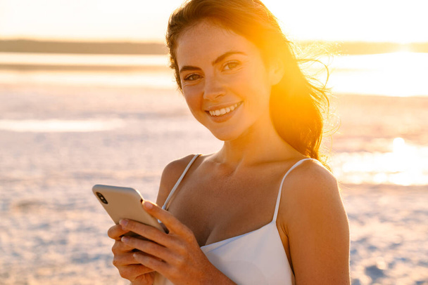 Image of a happy cheerful young pretty girl walking outdoors at the beach and using mobile phone - Foto, Bild