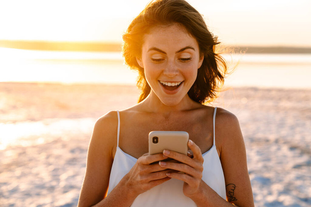Photo of a surprised young pretty girl walking outdoors at the beach and using mobile phone - Foto, afbeelding