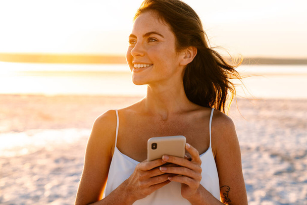 Image of a positive young pretty girl walking outdoors at the beach and using mobile phone - Valokuva, kuva