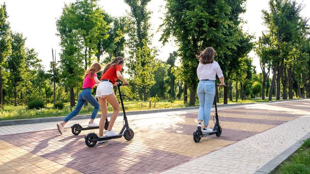 E-scooter, elektrische scooter, Ecologisch stedelijk vervoer, alternatieve energie, alternatieve levensstijl. Drie jonge meisje vrienden op vakantie met plezier rijden elektrische scooter door stadspark - Foto, afbeelding