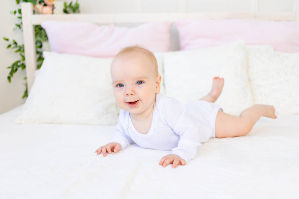 šťastný dítě dívka 6 měsíců v bílém obleku ležící na břiše na bílé posteli doma - Fotografie, Obrázek
