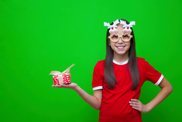 Wesoła dziewczyna w ciemnych włosach w czerwonym T-shircie i śmiesznych okularach trzyma pudełko z prezentem na Święta Bożego Narodzenia i Nowy Rok w rękach. - Zdjęcie, obraz