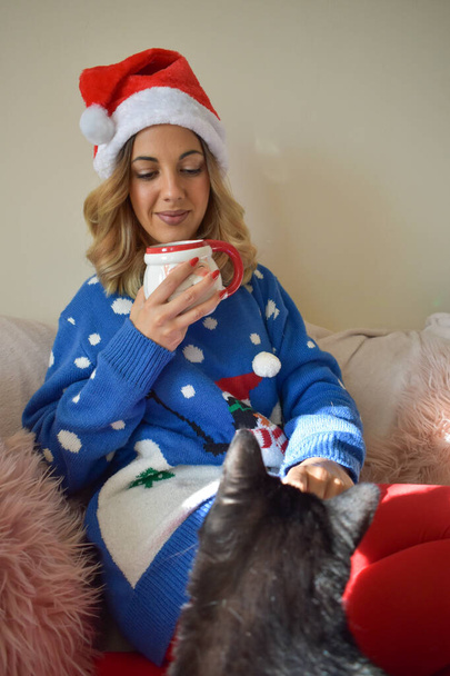 Girl dressed in Christmas clothes, having a coffee sitting on the sofa at home with her cat. - 写真・画像
