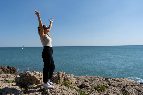 Pretty female celebrating and enjoying with raised up open arms standing on the rocks next to the sea.Freedom, happiness and people success lifestyle concept.Summer background.Copy space text. - Foto, afbeelding