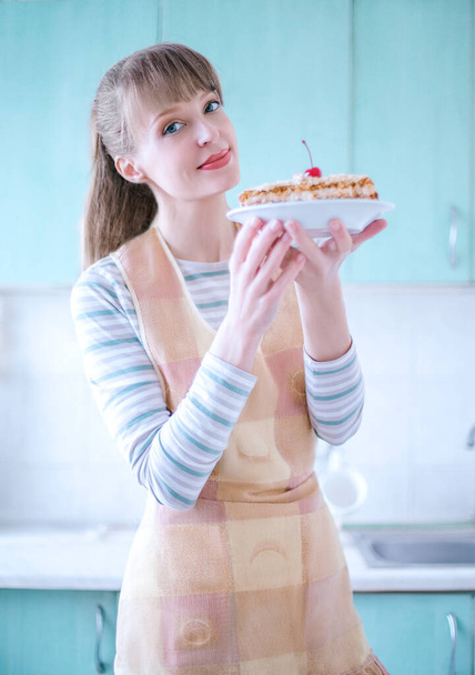 Woman is on his kitchen with a plate of homemade honey cake to present it for selling. European woman smiles, dressed with apron. Bright daylight picture. Vertical view. - Photo, Image
