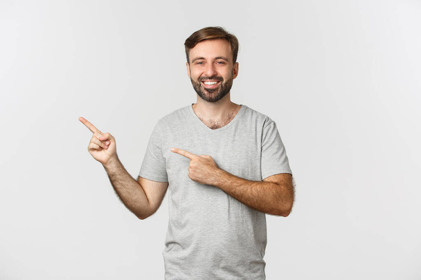 Imagem de cara feliz em roupas casuais sorrindo, apontando os dedos deixados para o seu logotipo ou promo, de pé sobre fundo branco - Foto, Imagem