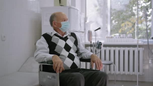 Emeklilik sağlık sorunları, tekerlekli sandalyede oturan yaşlı gri saçlı adamın portresi hastane içinde koruyucu maske takıyor. - Video, Çekim