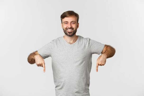 Retrato de hombre barbudo atractivo en camiseta gris, mostrando el logotipo, señalando los dedos hacia abajo, de pie sobre fondo blanco - Foto, imagen