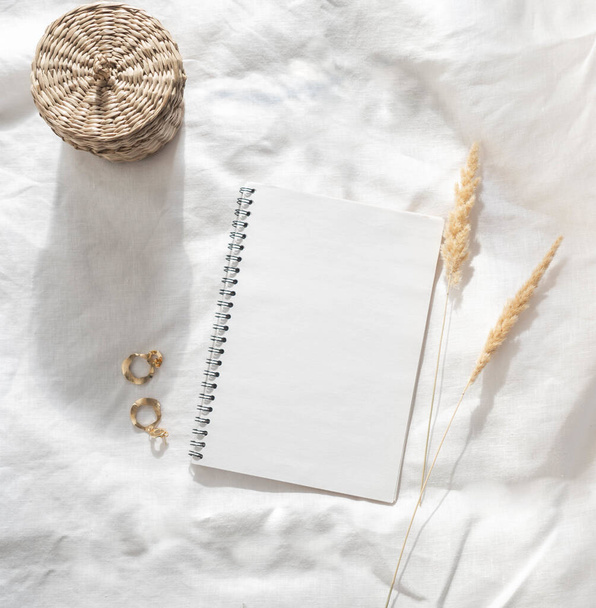 White blank notepad sheet with spiral, sprigs of reeds, gold earrings and wicker box on white crumpled textile. top view. Copy space. Flat lay - Photo, Image