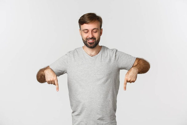 Atractivo modelo masculino caucásico en camiseta gris, apuntando y mirando hacia abajo, sonriendo complacido, de pie sobre fondo blanco - Foto, imagen