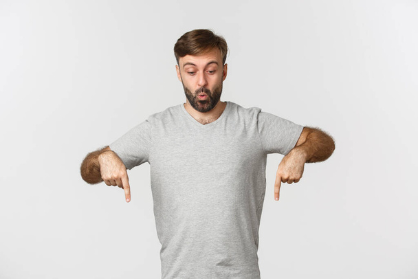 Surprised attractive guy in gray t-shirt, pointing and looking down fascinated, saying wow, standing over white background - Photo, Image