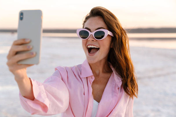 Image of a cute surprised shocked young girl in sunglasses taking a selfie by phone outdoors at the beach - Photo, Image