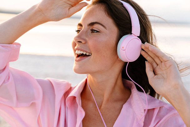 Image of a cheery pleased young girl in sunglasses listening music by headphones outdoors at the beach - Foto, Imagem