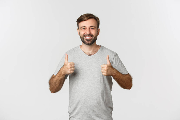 Imagen de hombre barbudo guapo en camiseta gris, mostrando pulgares hacia arriba en aprobación, como algo bueno, de pie sobre fondo blanco - Foto, Imagen