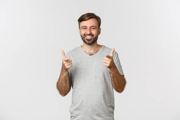 Image of satisfied smiling man in gray t-shirt, showing thumbs-up in approval, like something good, white background - Фото, изображение