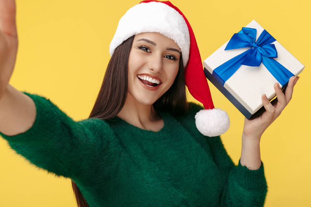 Fröhliche junge brünette Frau trägt Weihnachtsmann-Hut und grünen Pullover machen Selfie mit Geschenkbox über gelbem Hintergrund. - Foto, Bild