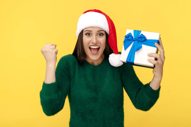 Excited young brunette woman wear santa claus hat and green pullover holding gift box over yellow background. - Foto, Imagem