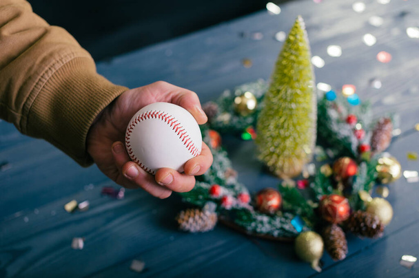 A man holds a baseball ball against the background with decoration on a wooden table. Festive composition for new year and christmas - Foto, Imagem