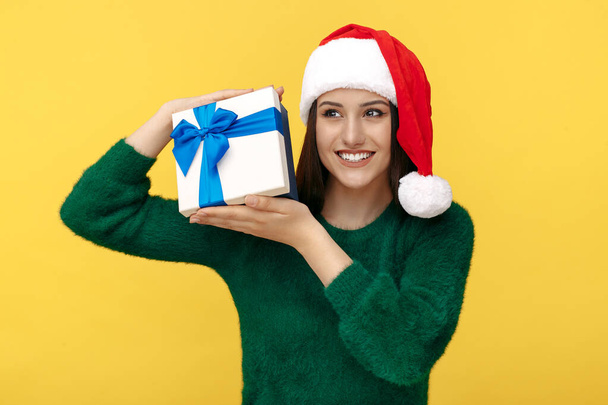 Smiling young brunette woman wear santa claus hat and green pullover holding gift box over yellow background. - Valokuva, kuva