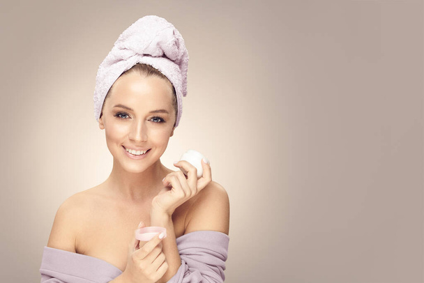 Beautiful smiling female model with clean skin, wearing in bathrobe and towel on head, hold cream on beige isolated with free copy space. - Foto, imagen