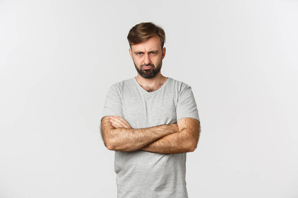 Portret van een beledigde knappe man, mokkend en boze grimas makend, handen over de borst defensief, staand over een witte achtergrond - Foto, afbeelding