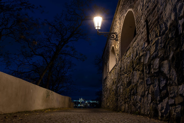 街路灯や15世紀の要塞の古い石壁からの光と、秋のチェコ共和国プラハの中心部で夜に歩行者のための地面に石を舗装 - 写真・画像