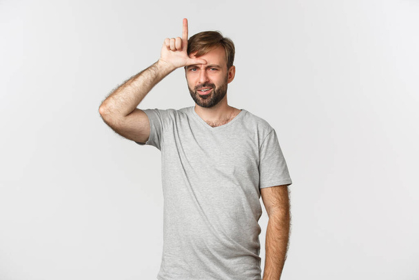 Πορτρέτο του αλαζόνα άνδρα σε γκρι t-shirt, χλευάζοντας πρόσωπο που έχασε, δείχνει χαμένος σημάδι στο μέτωπο και ψάχνει με απογοήτευση, στέκεται πάνω από το λευκό φόντο - Φωτογραφία, εικόνα
