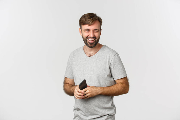 Imagem de homem barbudo bonito em camiseta cinza, segurando smartphone, olhando para a direita e rindo, de pé sobre fundo branco - Foto, Imagem
