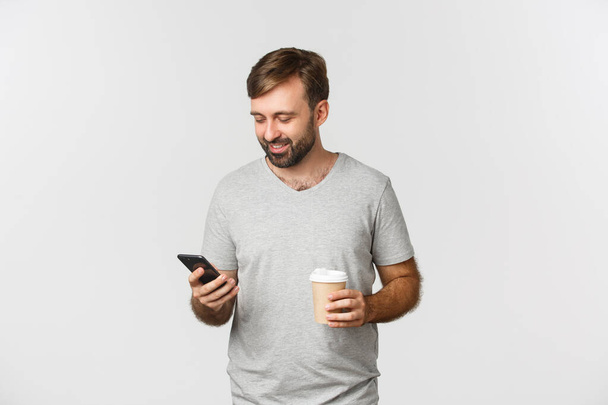 Imagen del hombre caucásico moderno con barba, bebiendo café y usando casualmente el teléfono móvil, de pie sobre fondo blanco - Foto, Imagen