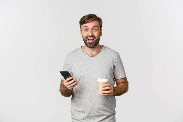 Imagem do homem caucasiano moderno com barba, bebendo café e casualmente usando telefone celular, de pé animado sobre fundo branco - Foto, Imagem