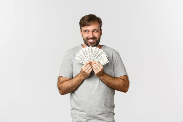 Chamtivý chamtivý muž s vousy, přemýšlející o nakupování, držení peněz a pohledu na levý horní roh, stojící nad bílým pozadím - Fotografie, Obrázek