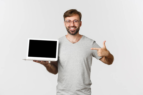 Cheerful bearded man in gray t-shirt, showing laptop screen and smiling, working from home, making thumbs-up in approval, standing over white background - Photo, image
