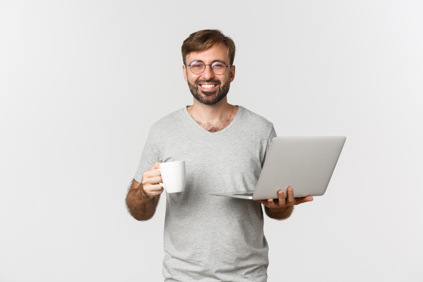 Jovem homem feliz trabalhando com laptop, bebendo café e sorrindo, de pé sobre fundo branco - Foto, Imagem