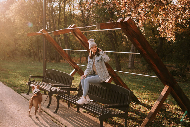 Uma jovem adolescente com um chapéu em um casaco de ganga sentado em um banco de parque e ao lado dela é um cão americano Staffordshire Terrier. Dia de outono na natureza - Foto, Imagem