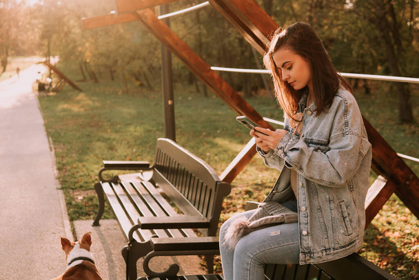 Вид збоку мила дівчина-підліток брюнетка, одягнена в джинси, сидить на лавці і використовує телефон. Осінні прогулянки в парку
 - Фото, зображення