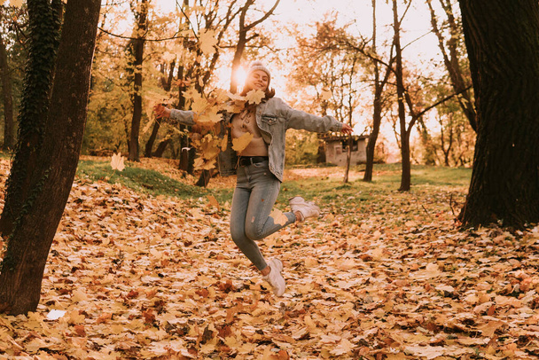 Kot pantolonlu şapkalı, yaprakları fırlatan ve parktaki havaya zıplayan şehirli genç bir kızın boyu. Ormanda güneşli bir sonbahar günü - Fotoğraf, Görsel