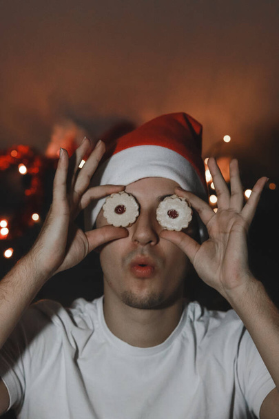 Дивний вираз молодого чоловіка з різдвяним капелюхом. Людина тримає різдвяне печиво замість очей. Печиво Лінц з полуничним варення замість очей. Комічні сірники. - Фото, зображення