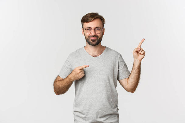 Wesoły brodaty mężczyzna uśmiecha się, wskazując palcami w prawym górnym rogu, pokazując logo, stojąc nad białym tłem - Zdjęcie, obraz