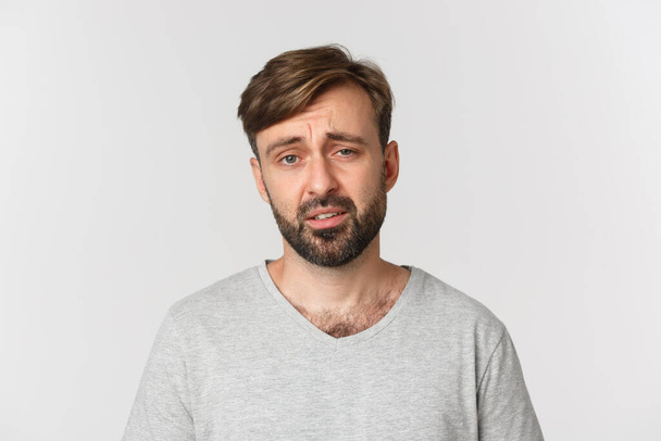 Homem barbudo cético e sem graça em camiseta cinza, franzindo a testa e olhando exausto, de pé sobre fundo branco - Foto, Imagem