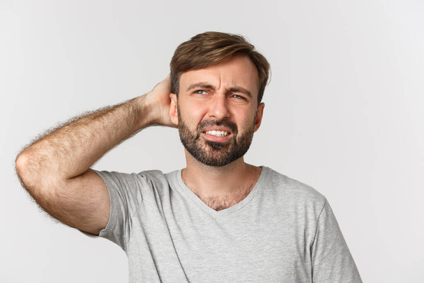Close-up of puzzled bearded man, looking confused at upper left corner, scartching head indecisive, standing over white background - Photo, Image