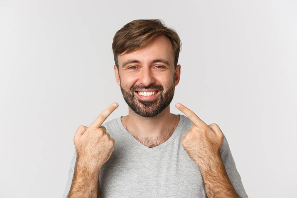 Detailní záběr pohledného usměvavého muže, který doporučuje zubní kliniku, ukazuje na své dokonalé bílé zuby, stojí nad bílým pozadím - Fotografie, Obrázek