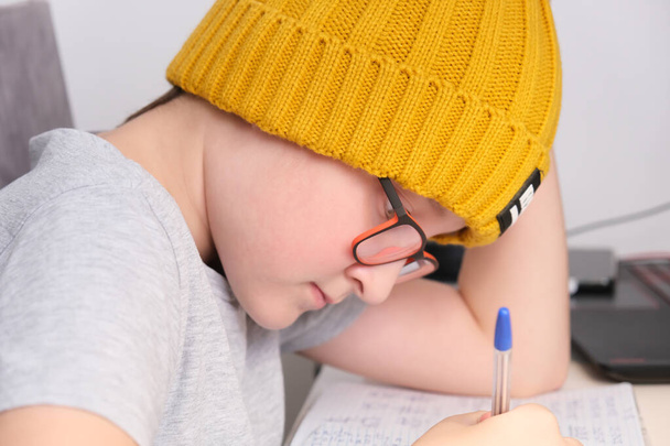 A boy in a trendy yellow knitted hat sitting by the table at home and doing homework. - Fotó, kép