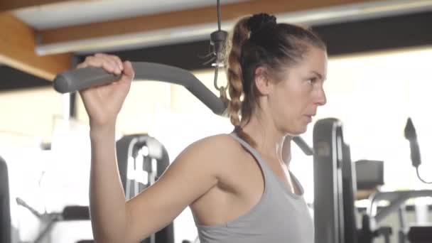 Piękna młoda kobieta w spodenkach sportowych i szarej koszulce z butli zaangażowana w fitness w siłowni ściska dłonie na specjalnym symulatorze - Materiał filmowy, wideo