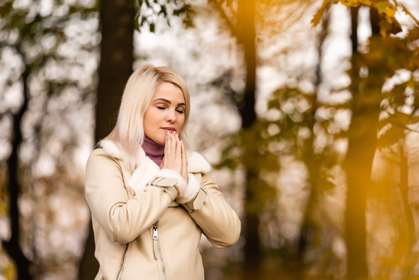 Νεαρή γυναίκα με σφιγμένα χέρια, προσεύχεται προσευχή, στέκεται στη φύση το φθινόπωρο - Φωτογραφία, εικόνα