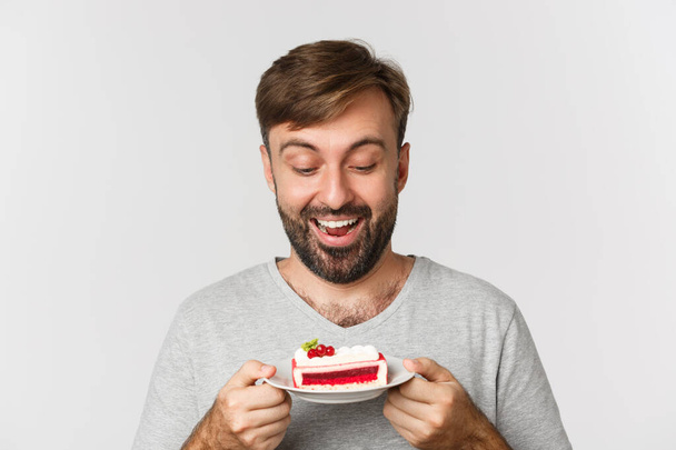 Close-up van knappe glimlachende man met taart, staande over witte achtergrond, opgewonden om dessert te eten - Foto, afbeelding