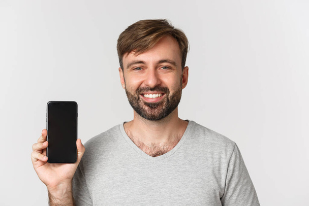 Primer plano de hombre guapo sonriente en camiseta gris, mostrando la pantalla del teléfono móvil, recomendando aplicación o sitio de compras, de pie sobre fondo blanco - Foto, imagen