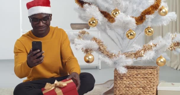 Hombre negro afroamericano con gafas se sienta en el suelo de la casa cerca del árbol de Navidad lleva sombrero de Santa Claus sostiene teléfono inteligente habla a la cámara del teléfono móvil hace video para el blog se comunica en el chat en línea - Imágenes, Vídeo