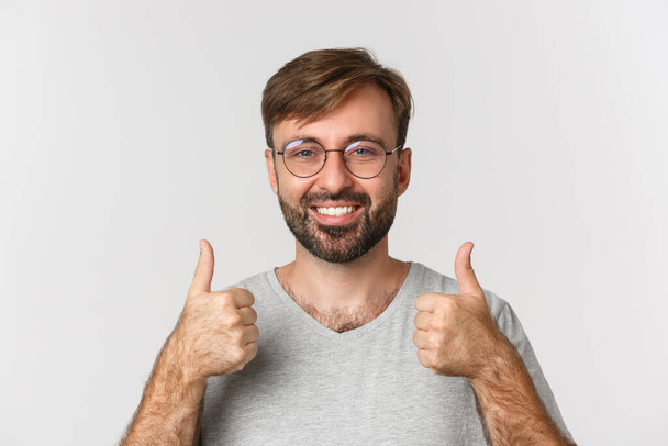 Primer plano del alegre hombre caucásico con barba, con gafas y camiseta casual, sonriendo y mostrando los pulgares hacia arriba en aprobación, recomiendan el producto, de pie sobre fondo blanco - Foto, imagen