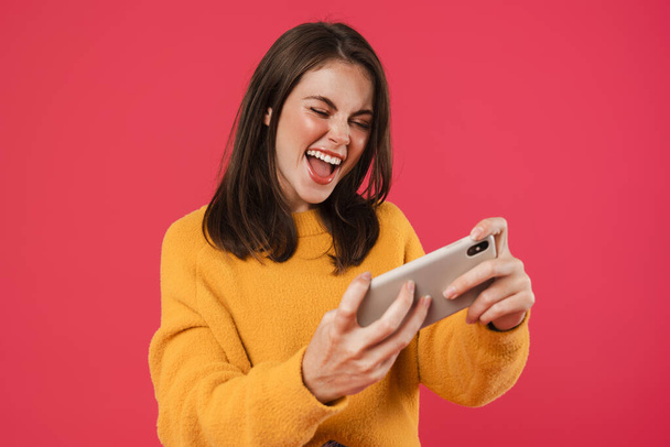 Image of excited brunette girl playing video game on cellphone isolated over pink background - Zdjęcie, obraz